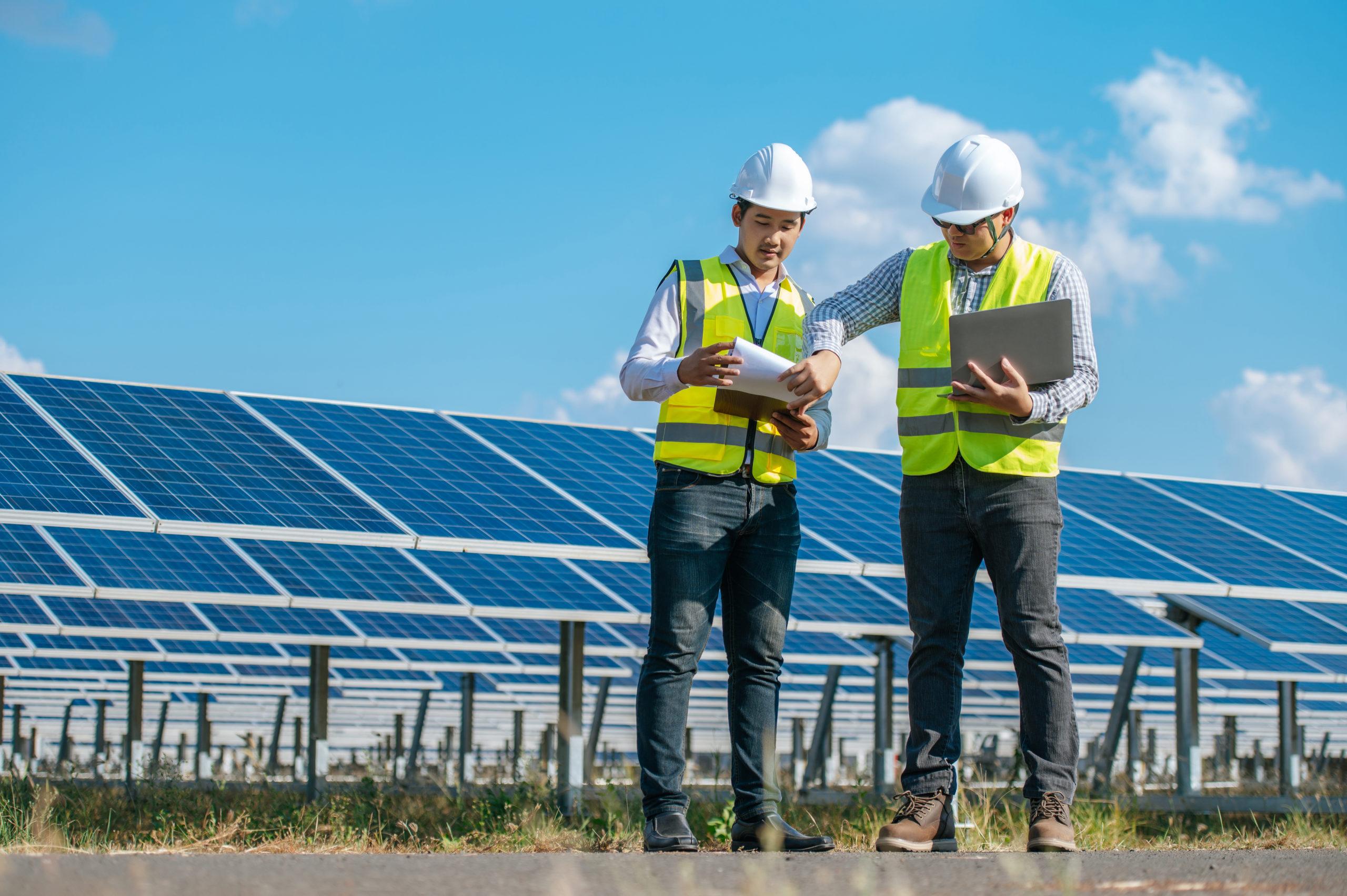 Two Asian young engineers walking along rows of photovoltaic panels in solar farm, They use  laptop computer and talking together